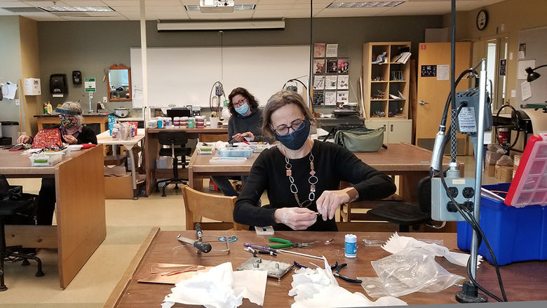 Three students wearing masks sitting at work tables and making jewelry.