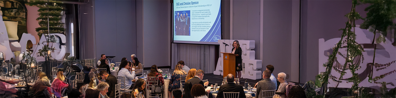 A woman speaks at a podium in front of a seated audience during a formal event, with a large presentation screen behind her displaying information about a scholarship.