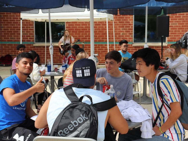 Students sit at an outdoor table under blue umbrellas, eating and talking, with a guitarist performing on a small stage nearby.