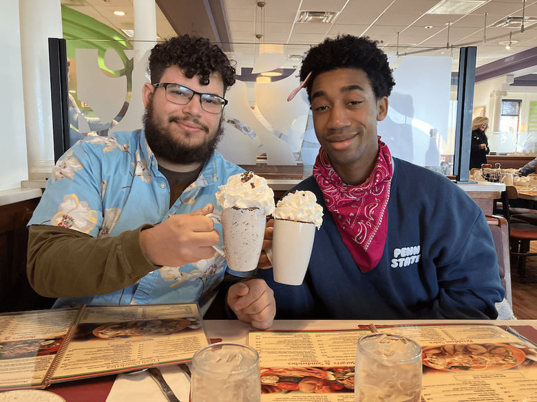 Two people at a restaurant table hold up milkshakes topped with whipped cream, with menus and water glasses in front of them.