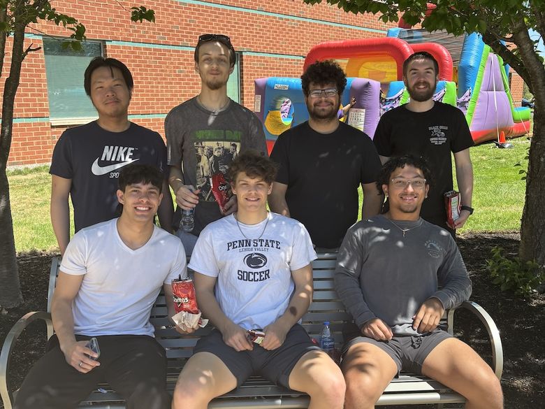 Six people pose outdoors near a bench under trees, with a brick building and inflatable bounce house in the background.