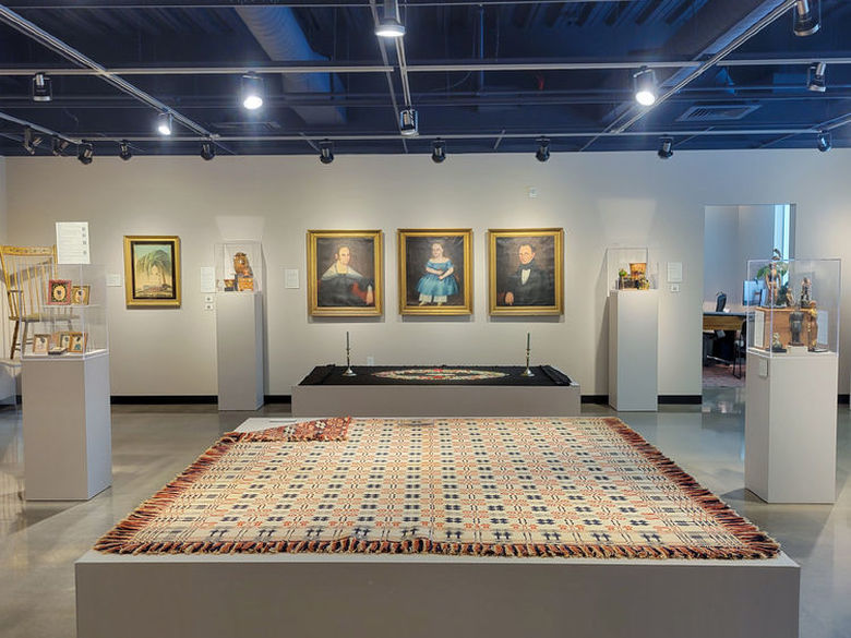 Interior of the Ron K De Long Gallery featuring a large patterned rug on a platform, framed portraits on the wall, and artifacts in glass cases.