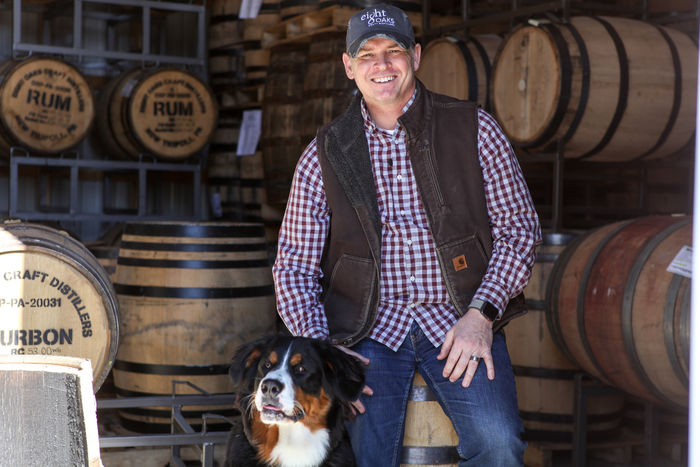 photo of man in vest with dog surrounded by barrels