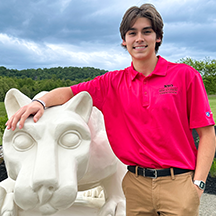 PSU-LV male student in bright pink New Student Orientation leader shirt smiling, standing next to the PSU-LV lion shrine.