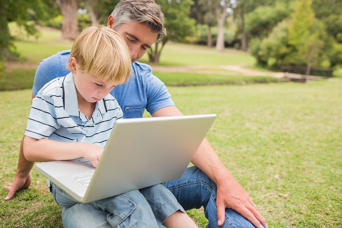 Father and young son sitting outside on the lawn., looking at laptop.