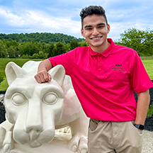 PSU-LV male student in bright pink New Student Orientation leader shirt smiling, standing next to the PSU-LV lion shrine.