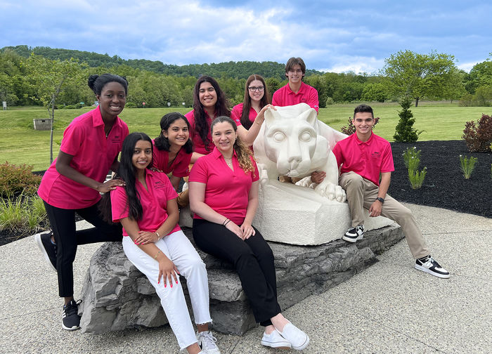 8 Penn State students in pink New Student Orientation shirts are gathered around the Penn State Lion Shrine statue.