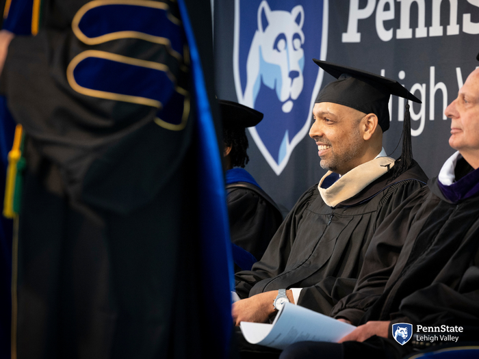 a man in black commencement regalia smiles as he sits on stage 