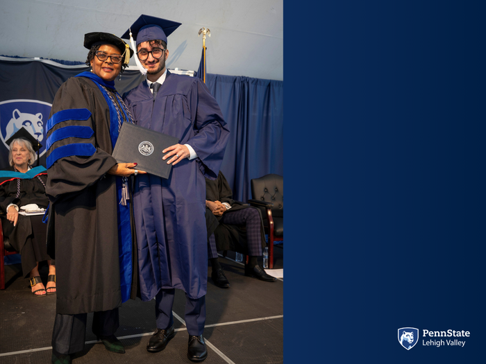 Two people in regalia pose with a diploma on a commencement stage