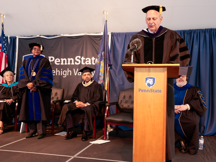 a man in black regalia stands at a podium at a commencement ceremony