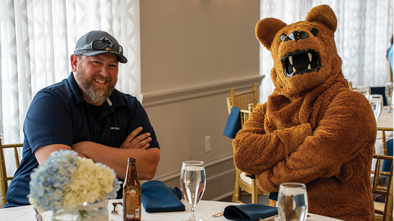 Male golf player and Nittany Lion mascot sitting at a table smiling at the camera.