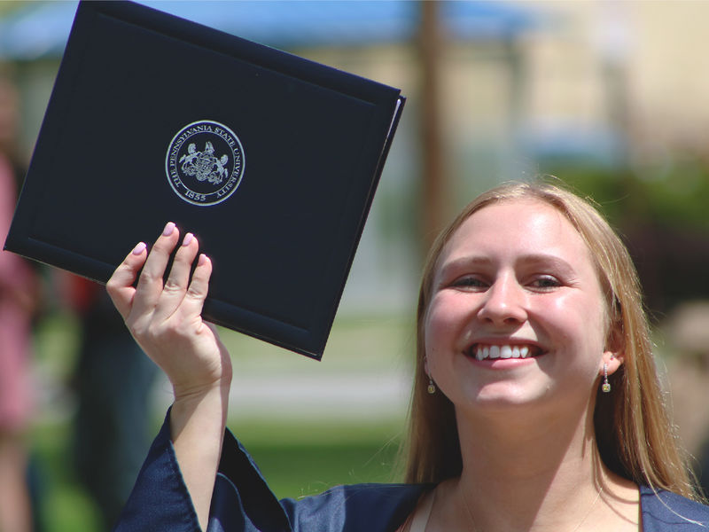Penn State graduate holds up diploma and smiles while outdoors