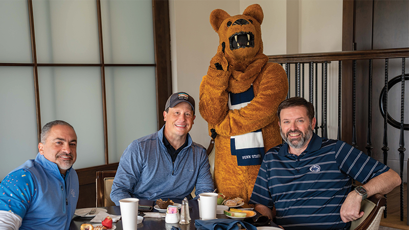 Three male golfers sitting in front of the Nittany Lion mascot.