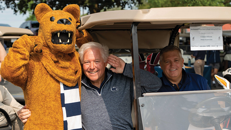 Nittany Lion mascot standing next to two male golfers sitting in a golf cart. 
