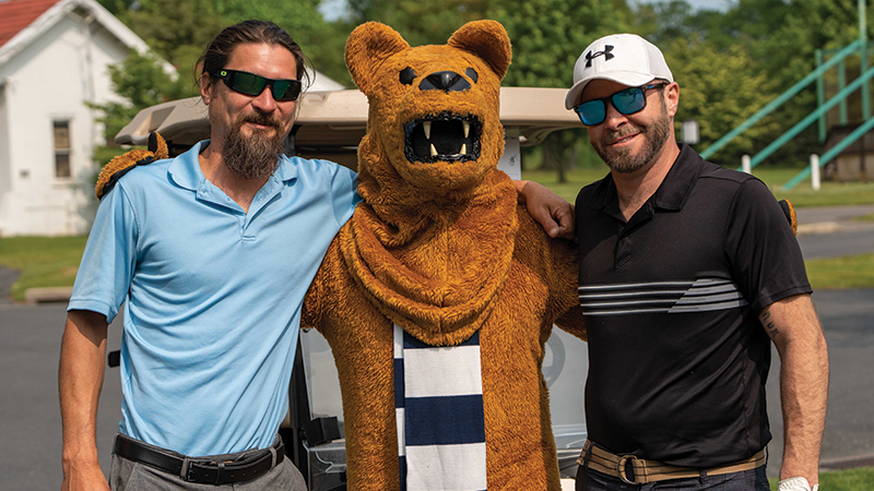 Two male golfers stand arm in arm with the Nittany Lion mascot.