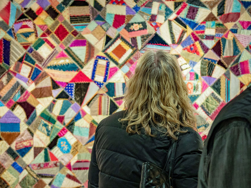 A woman stands in front of a patchwork quilt composed of brightly-colored, patterned fabrics.