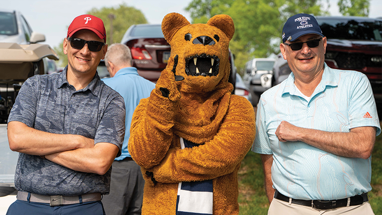 Two male golfers stand on either side of the Nittany Lion mascot
