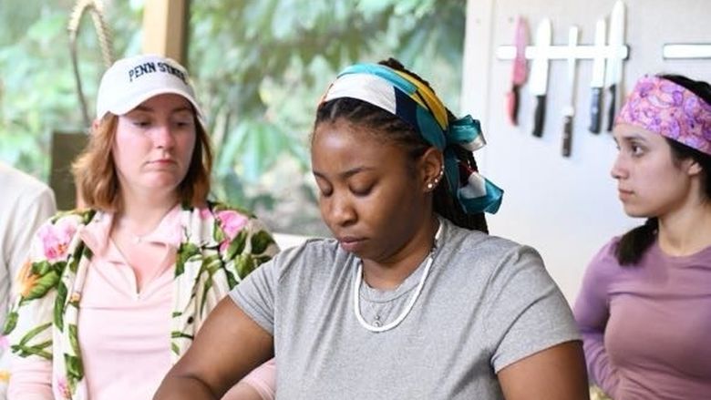 A student wearing a gray shirt and colorful headband prepares food in a kitchen
