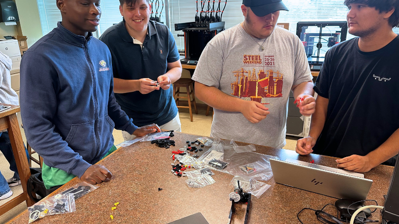 male college students collaborate on an engineering project at a long desk in a classroom