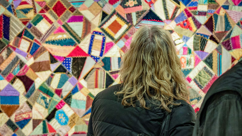 A woman stands in front of a patchwork quilt composed of brightly-colored, patterned fabrics.