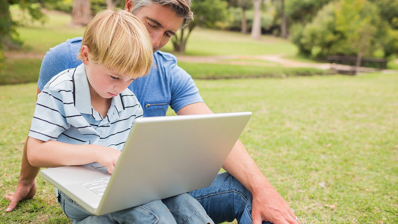 Father and young son sitting outside on the lawn., looking at laptop.