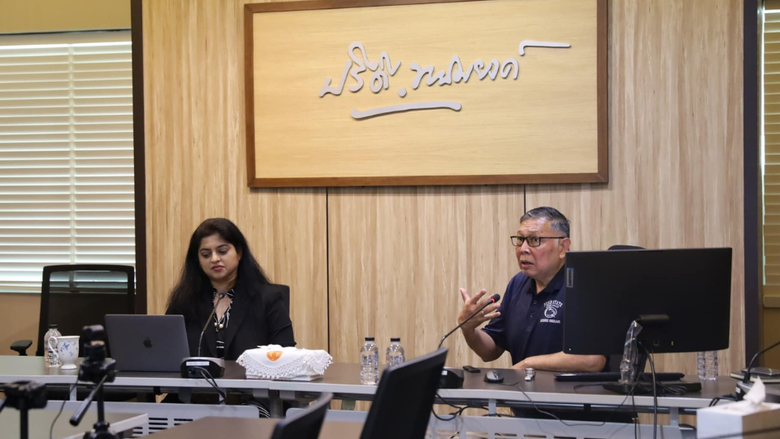 A man speaks to a group of people while sitting next to a woman behind a long desk