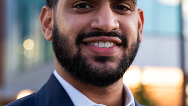 Man wearing a blue suit jacked and blue shirt looking at camera and smiling. He is standing outside in front of buildings.