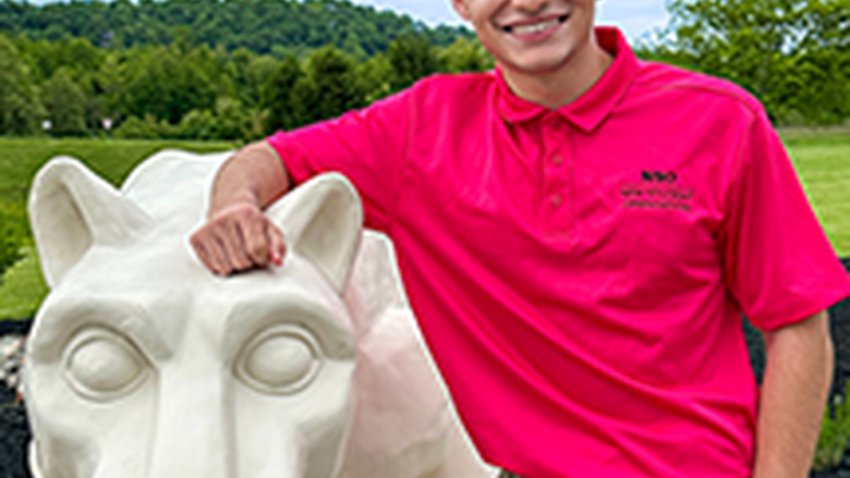 PSU-LV male student in bright pink New Student Orientation leader shirt smiling, standing next to the PSU-LV lion shrine.
