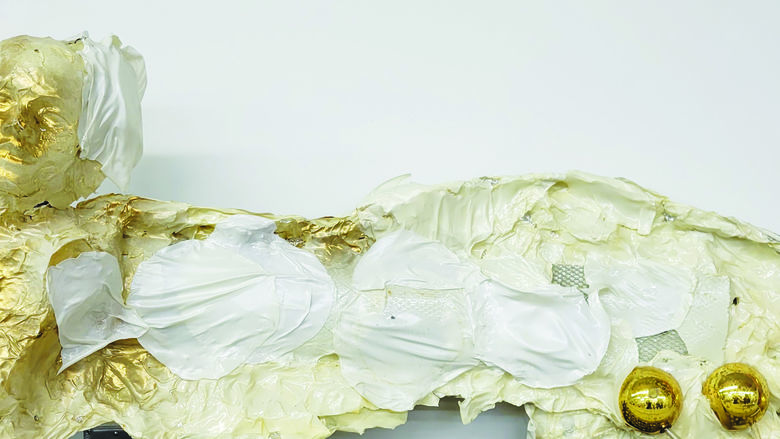 sculpture of human with extended wing in place of an arm. Figure wrapped in various fabrics, painted white and then accented with gold. Embedded in outer edge of wing are several gold balls 