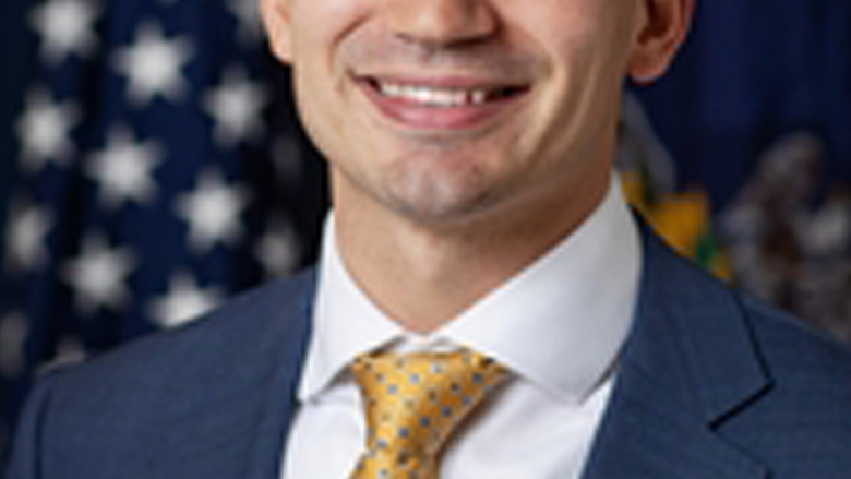 Man sitting in front of an American flag smiles at camera. He is wearing a navy suit, whit shirt, and yellow tie.