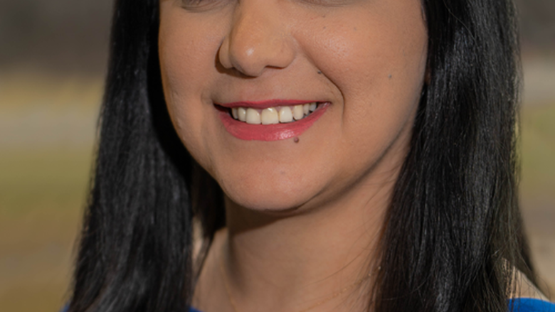 Woman with long dark brown hair,wearing a bright blue top smiles at the camera.