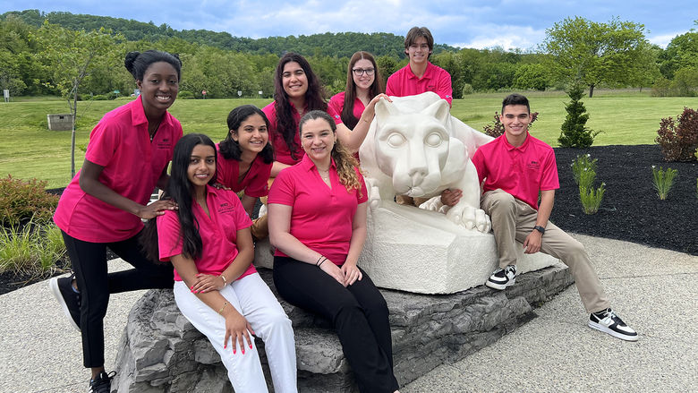 8 Penn State students in pink New Student Orientation shirts are gathered around the Penn State Lion Shrine statue.
