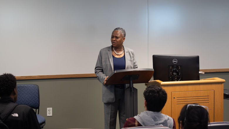 woman in blazer standing behind lectern speaking to class