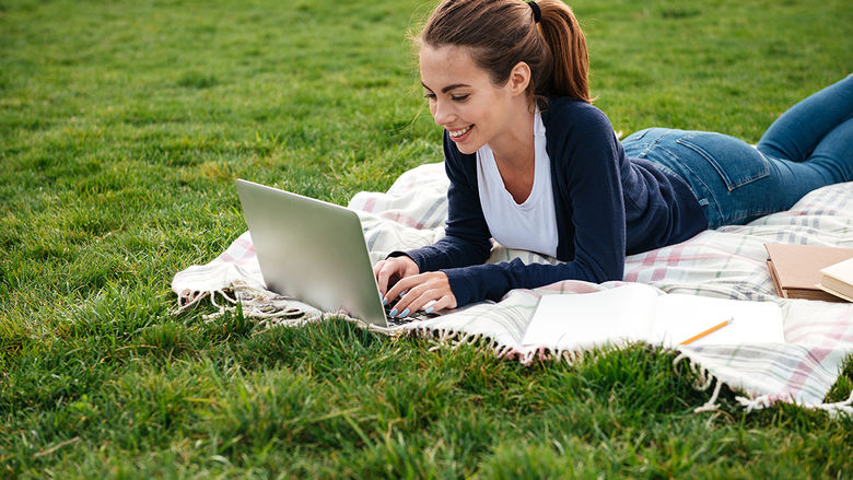 Happy teenage girl laying on blanket outside on the grass, typing on a laptop.
