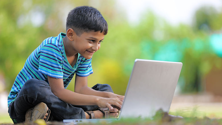 A happy young boy sitting cross-egged on the grass outside looking at the laptop computer