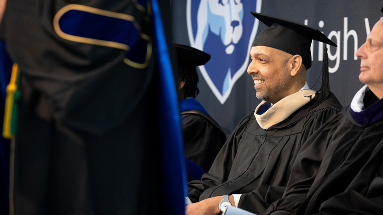 a man in black commencement regalia smiles as he sits on stage 