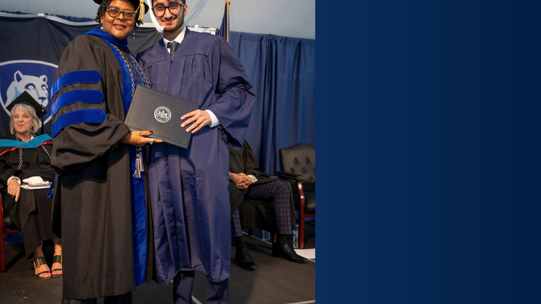 Two people in regalia pose with a diploma on a commencement stage