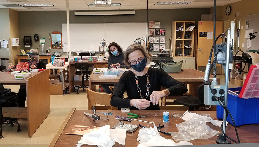 Three students wearing masks sitting at work tables and making jewelry.